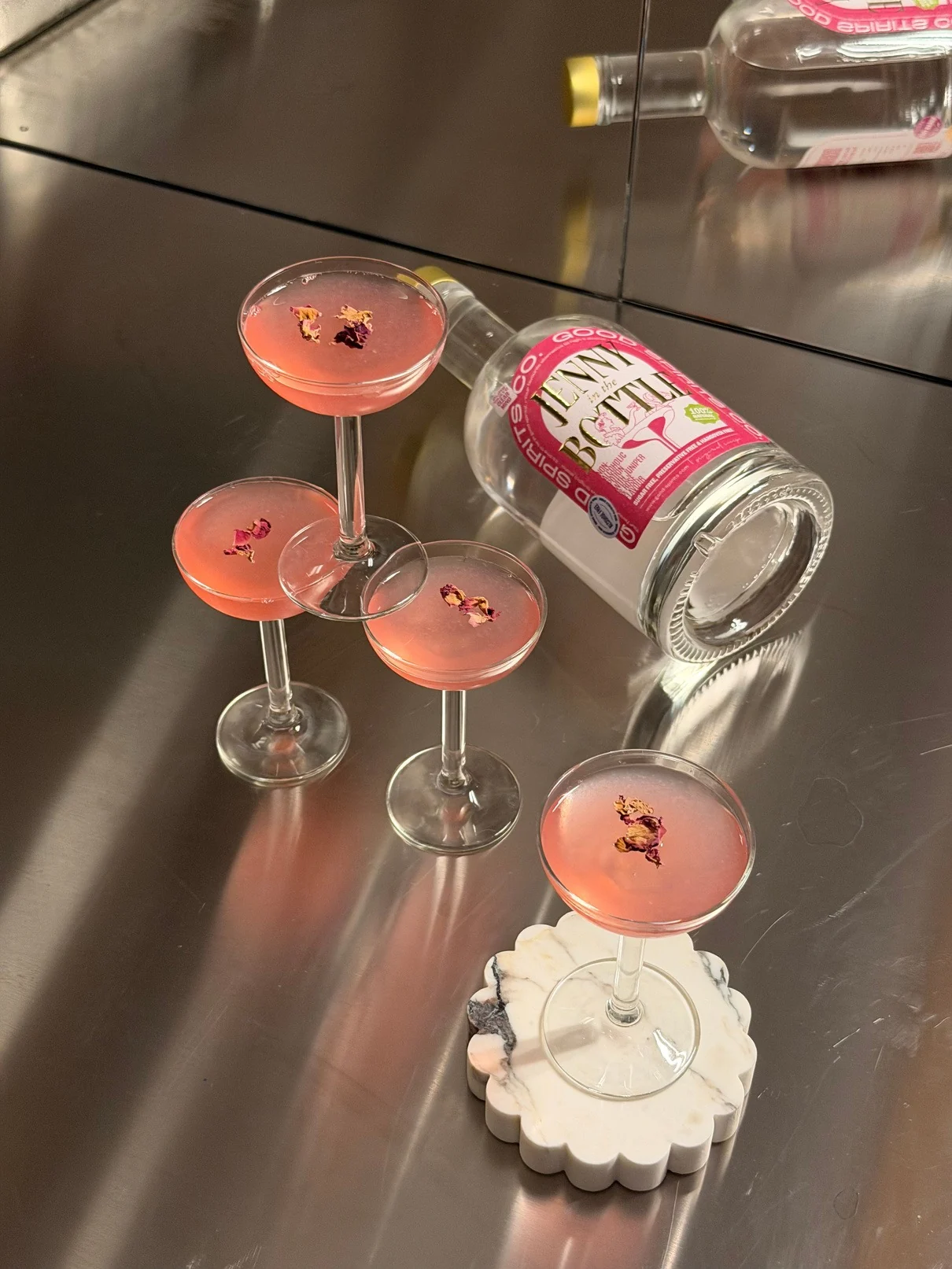 Four rose-topped coupe glasses beside a bottle of Jenny in the Bottle Rosé